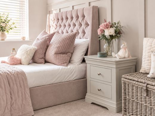 Toulouse Grey Painted 2 Drawer Large Assembled Bedside Table beside a blush upholstered bed and wicker storage chest