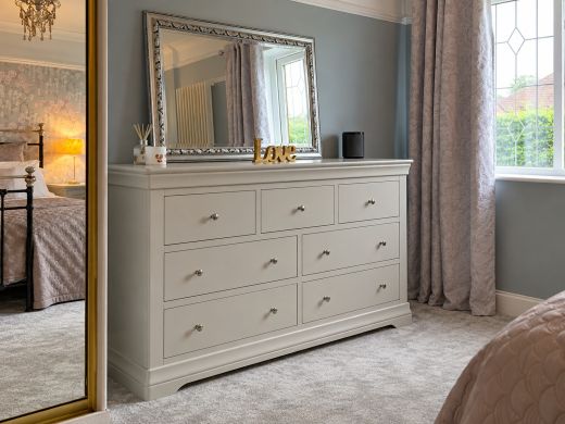 Toulouse Grey Grande extra large 3 over 4 chest of drawers with chrome knobs and a silver mirror, beside a curtained bedroom window