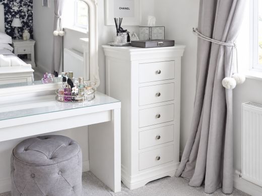 Toulouse white painted five-drawer Wellington tallboy beside a dressing table and ornate mirror in a bright bedroom with grey curtains