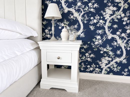 Toulouse white painted one-drawer bedside table with an open shelf, pleated lamp and candle under a glass cloche against navy floral wallpaper
