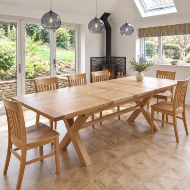 Provence 2.8m double extending cross-leg oak dining table with square corners and oak slatted chairs in a bright dining room with glass pendants and a wood-burning stove